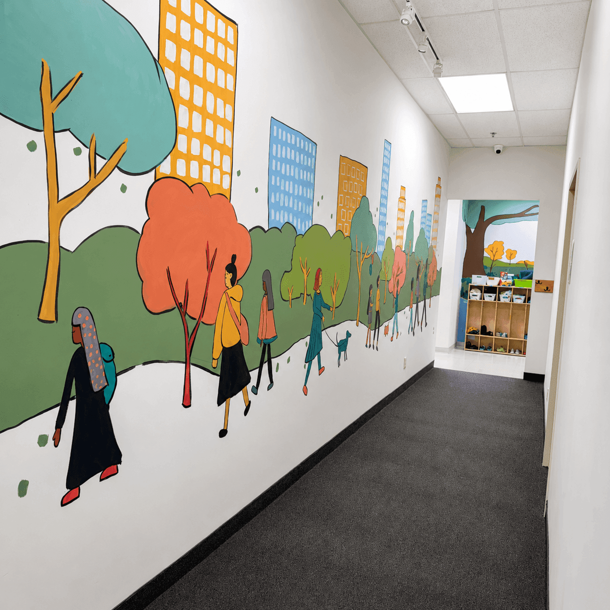 A long hallway painted with a colorful mural of people walking through a park toward an open room
